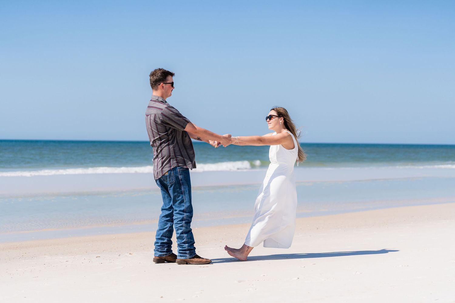 Couple holding hands and dancing on a sunny white sand beach with calm ocean waves in the background.