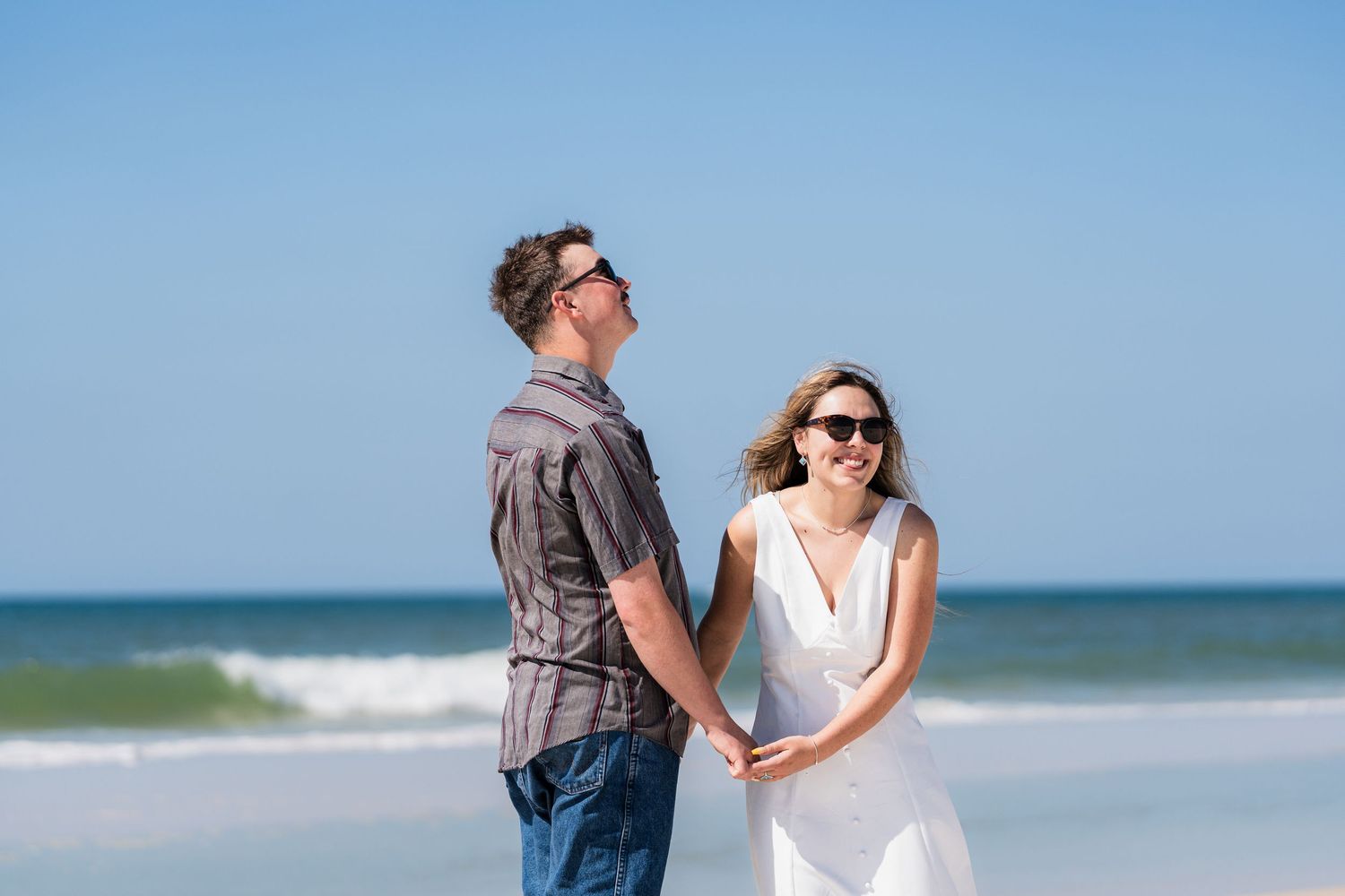Happy couple holding hands on a sunny beach, woman in white dress and man in gray shirt, both wearing sunglasses.