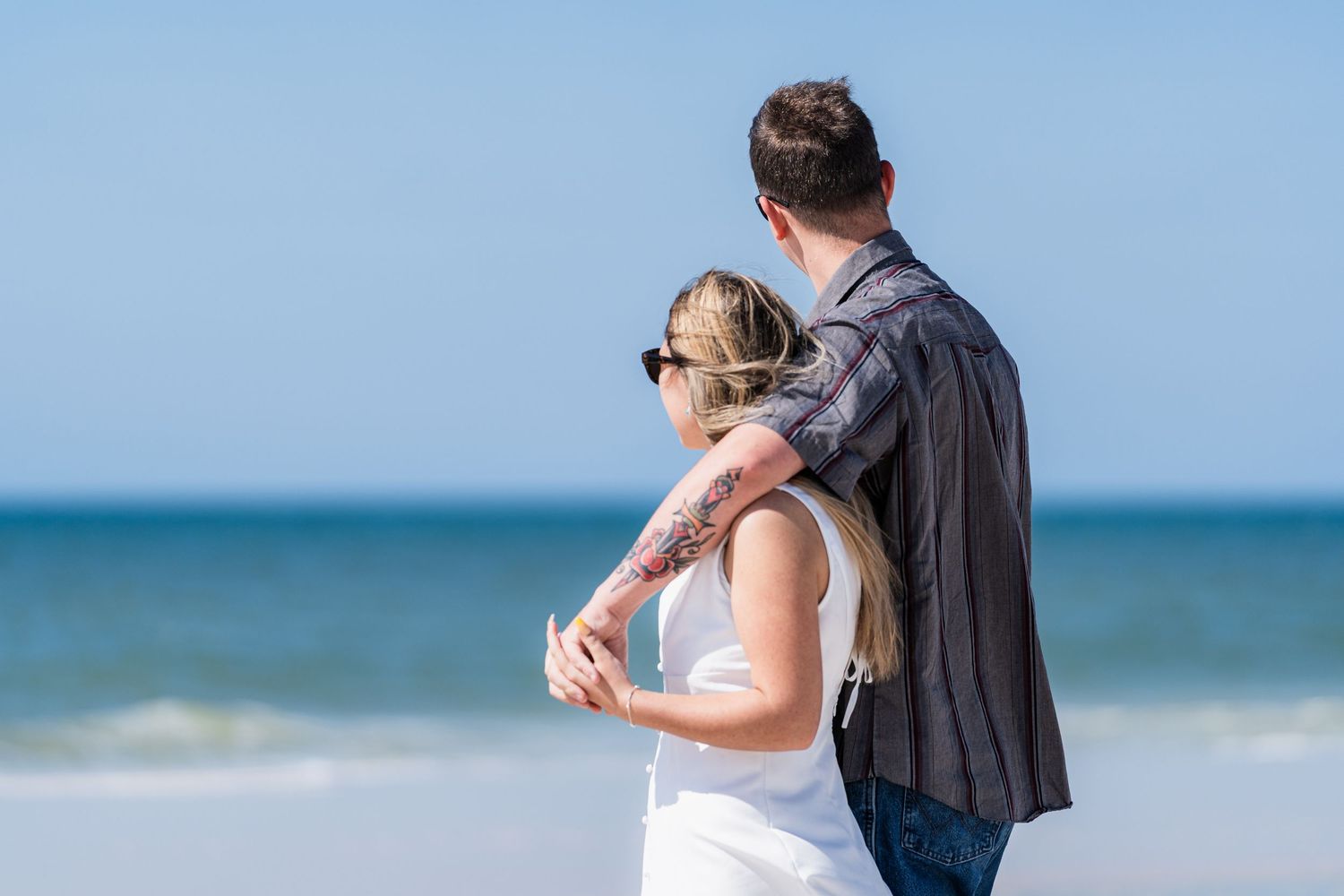 Couple embracing on a sunny beach, gazing at the ocean waves, woman with tattoos wearing sunglasses.
