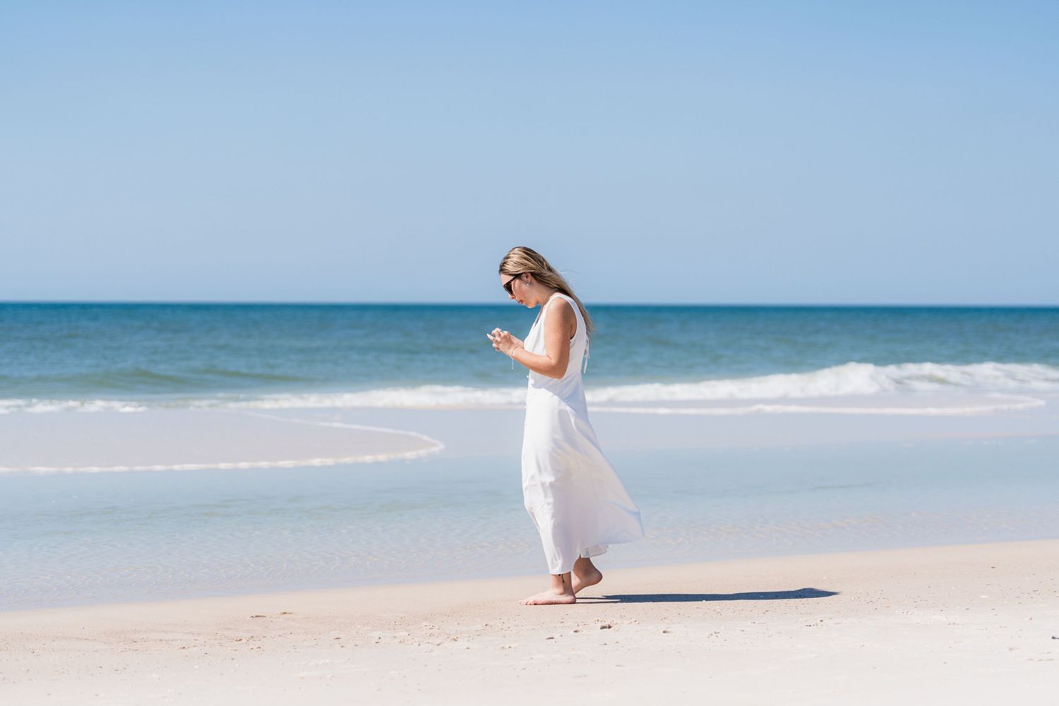 Woman in white dress and sunglasses walking barefoot on a sunny white sand beach with blue ocean waves.