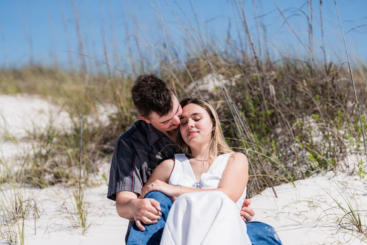 Romantic couple sharing a tender moment on white sand dunes with beach grass on a sunny day.