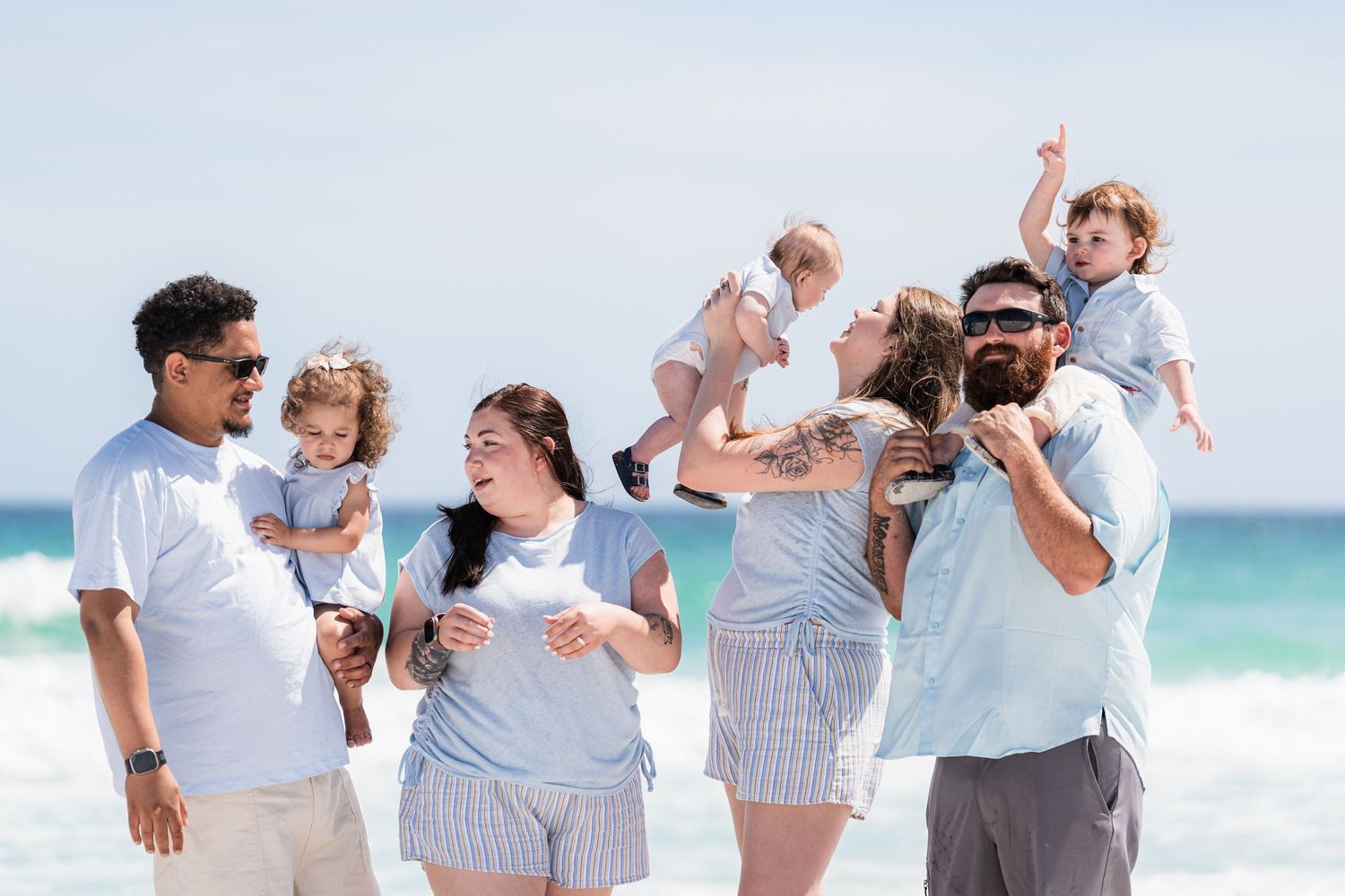 Family in matching blue outfits enjoying a sunny beach day, laughing and lifting children in the air.