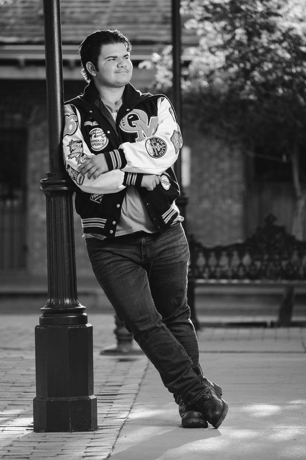 Black and white portrait of someone posing against a lamp post wearing a varsity jacket with patches.
