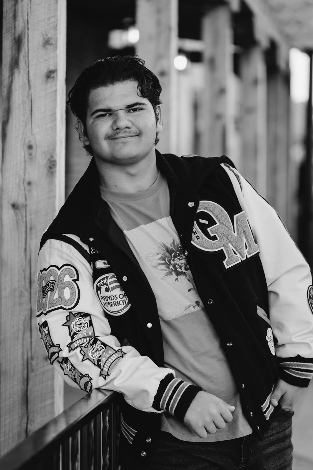 Black and white portrait of student wearing a varsity letterman jacket with patches leaning against wooden post.