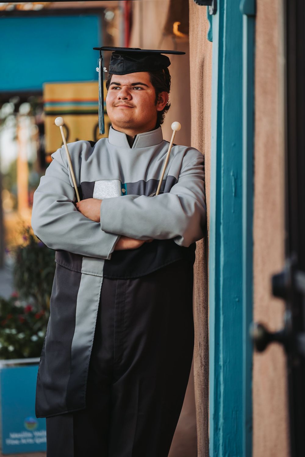 A person wearing a gray and black uniform stands with crossed arms against a teal-colored wall in an outdoor setting.