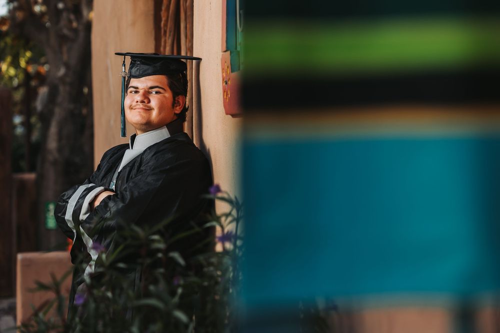 A person in graduation attire and cap smiles while posing for a photo in front of a blurred background.
