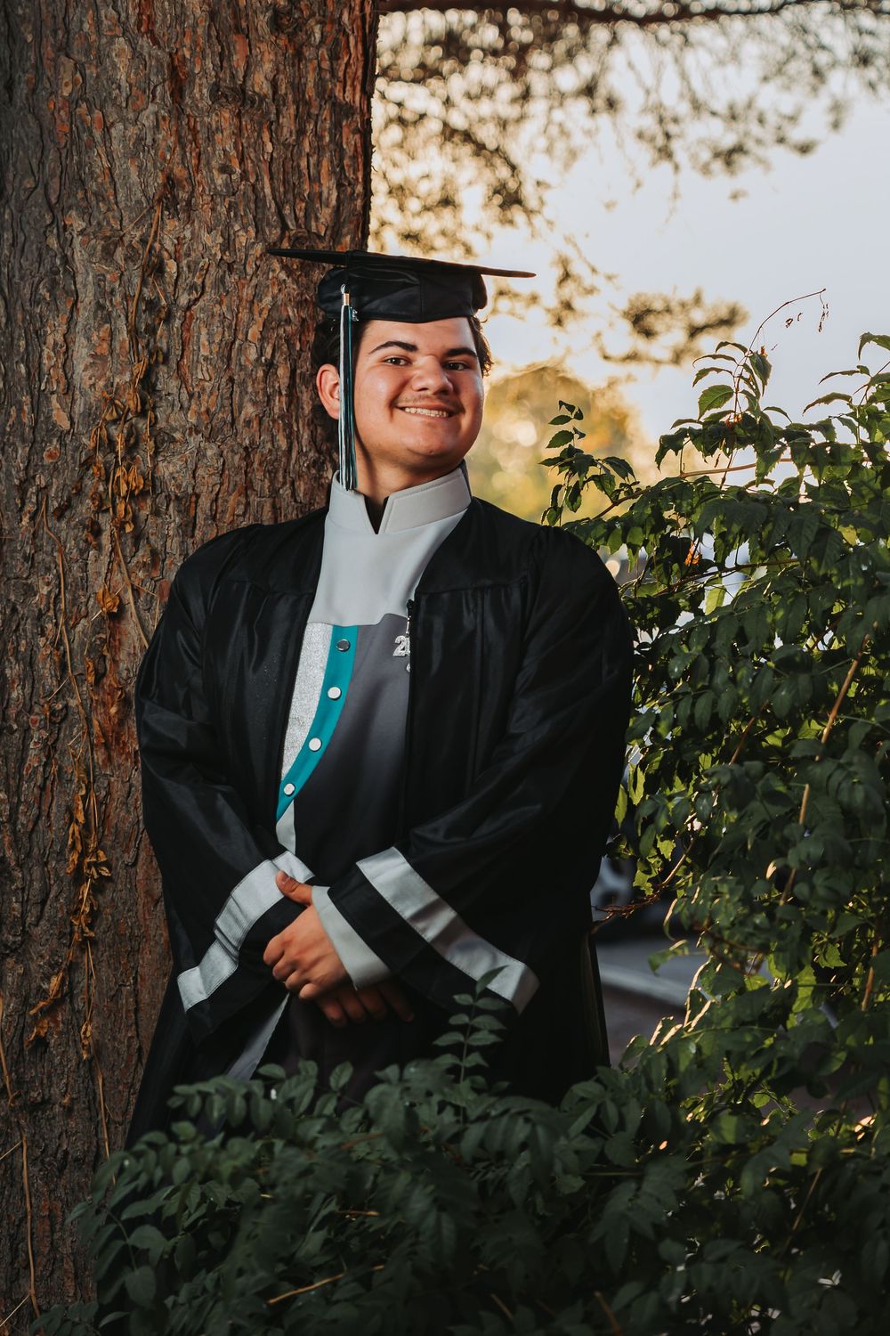 A graduate in academic regalia poses outdoors next to a tree trunk during golden hour lighting.