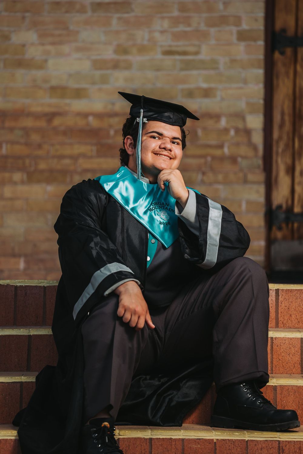 Graduate in cap and gown sits on brick steps wearing a teal sash while posing for graduation photos.