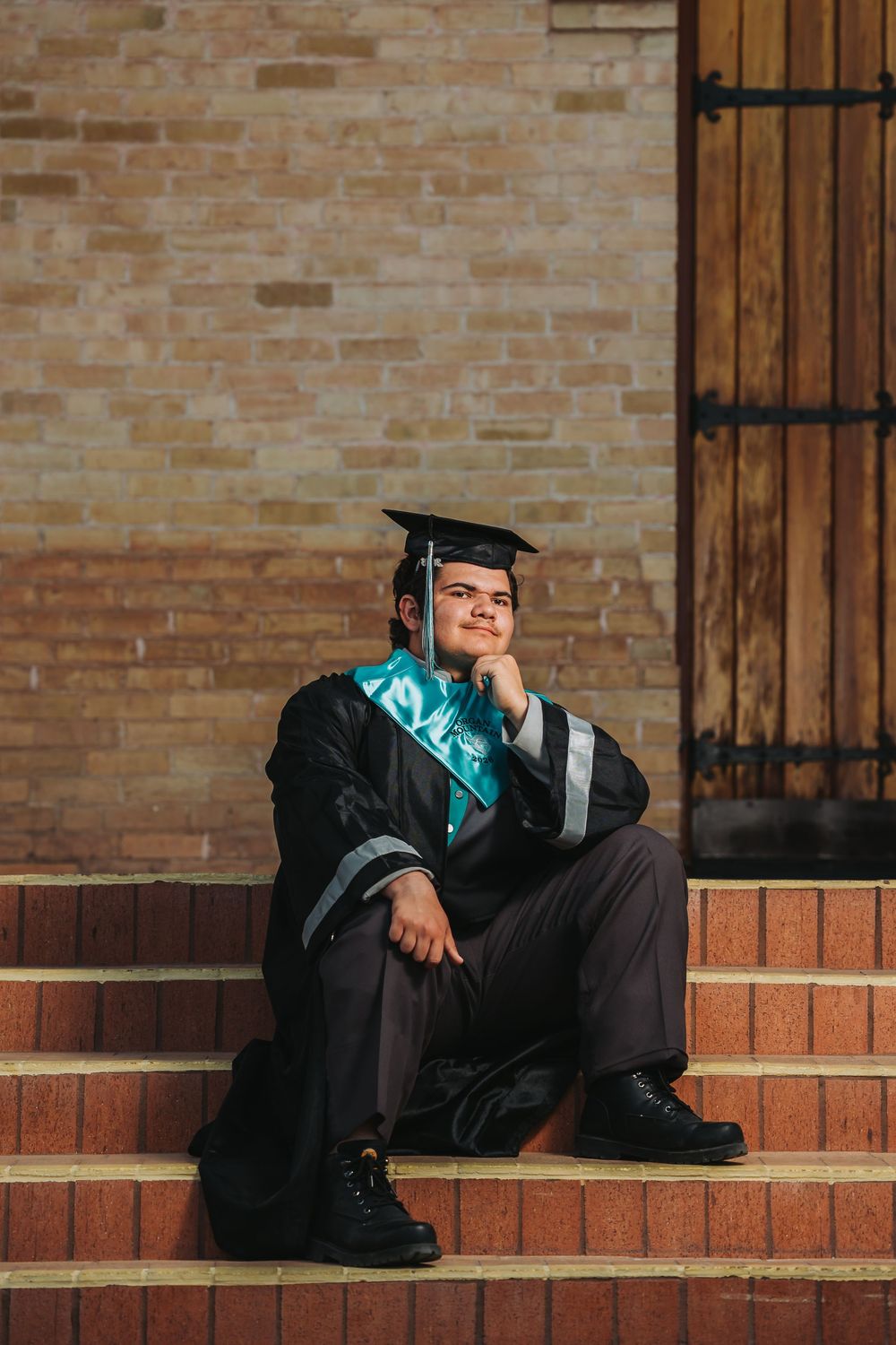 Graduate in cap and gown sits thoughtfully on brick steps during graduation photoshoot.