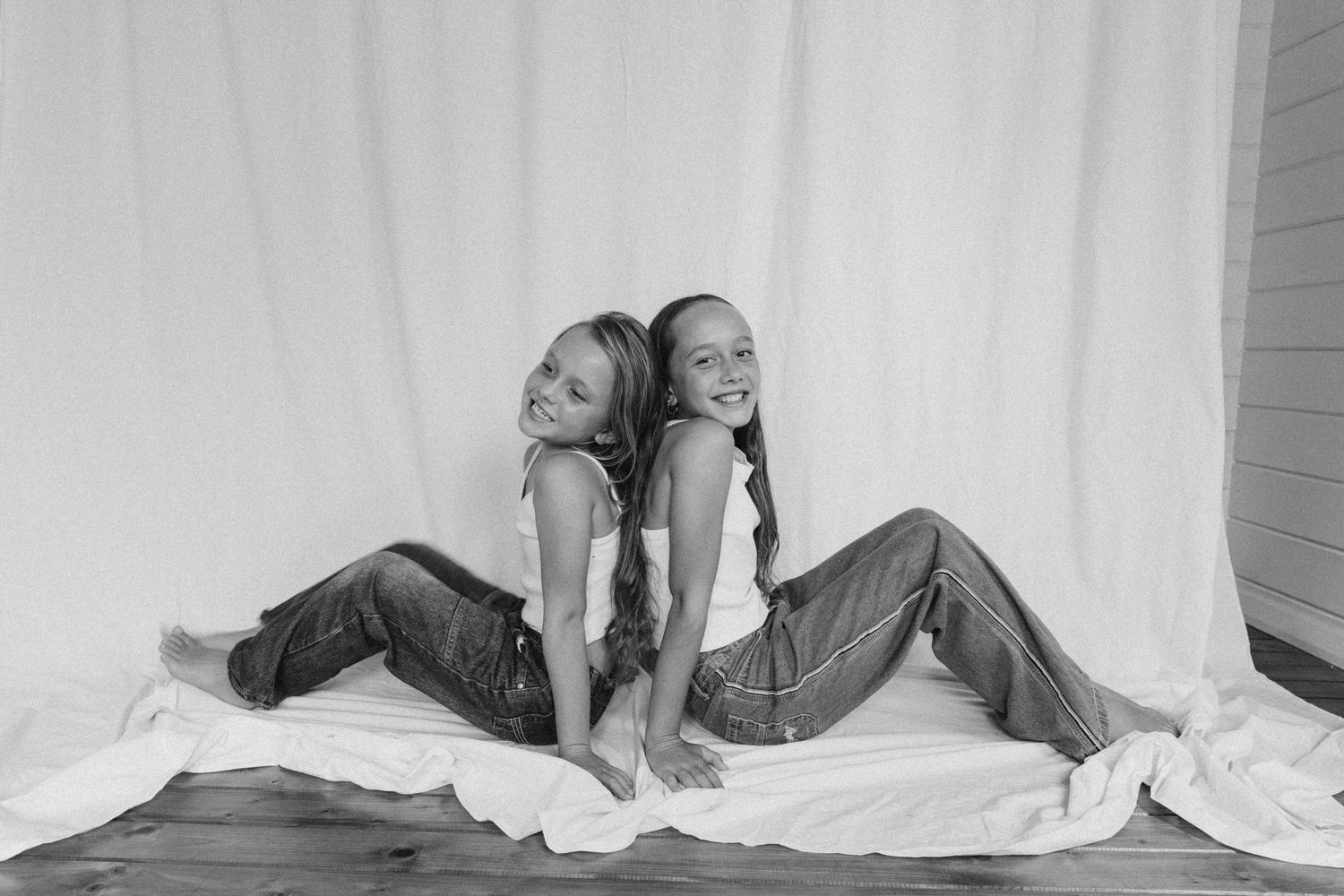 Black and white photo of two girls in white tops and jeans sitting back to back smiling on white fabric in New Zealand.