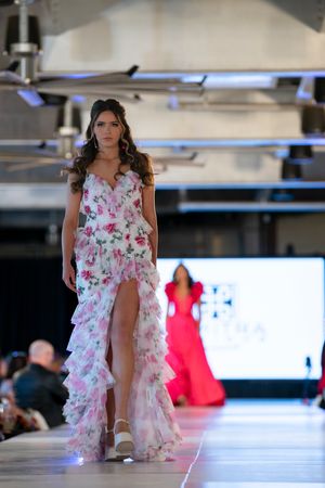 Model walks runway in flowing white floral dress with ruffled details at fashion show.