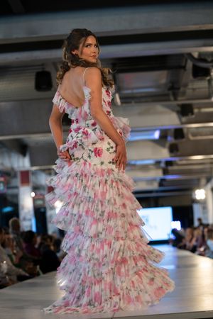 Fashion model wearing a white floral tiered ruffle gown with pink accents on a runway.