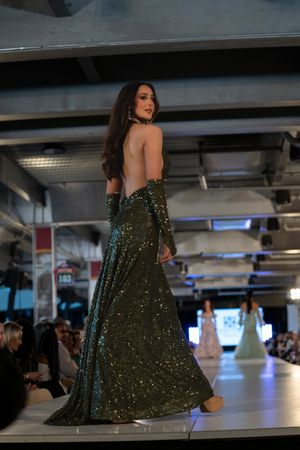 Model walks runway in sparkling dark green backless evening gown at fashion show.