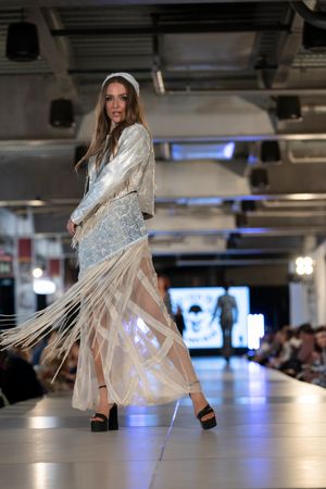 Flowing white dress with fringe details being showcased on illuminated runway.