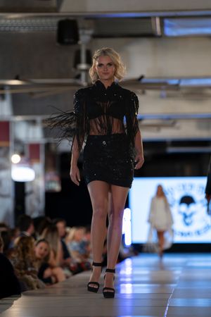 Model showcases edgy black fringe crop top and mini skirt ensemble on illuminated runway.