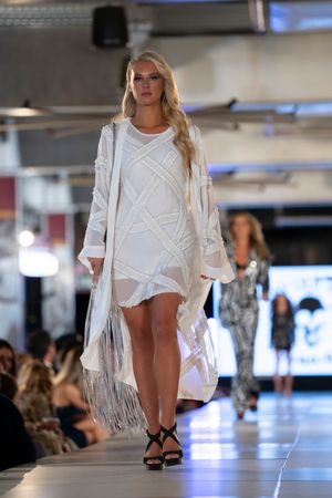 Fashion model walks runway in white summer ensemble with fringe details at indoor fashion show.