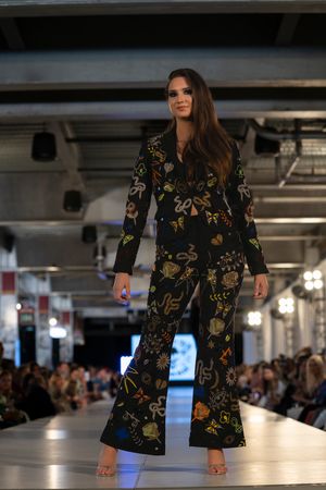 Model presents black floral print dress on runway with industrial ceiling visible.