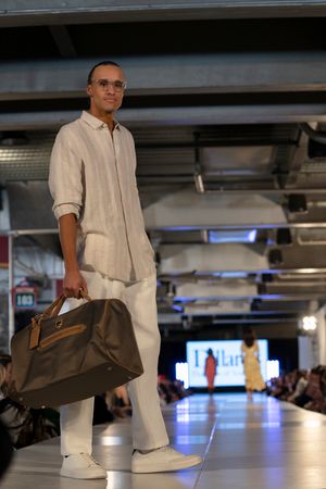 Model showcases all-white casual wear collection with brown accessories on illuminated catwalk.