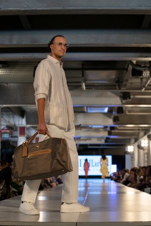 Model showcases casual menswear with brown leather bag on illuminated runway at fashion show.