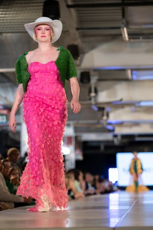 Fashion model walks runway in bright pink ruffled dress with green puff sleeves and white sunhat during fashion show.