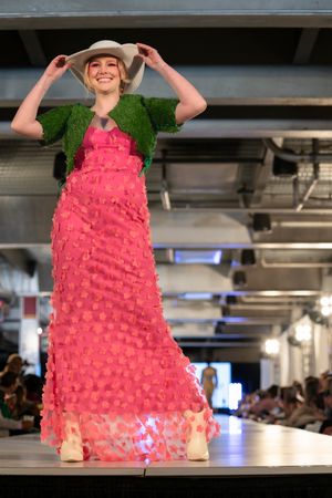 Model walks runway in bright pink ruffled maxi dress with green shrug and beige hat at fashion show.
