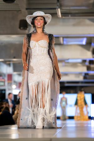 White lace dress with fringe details and wide-brimmed hat on fashion runway.