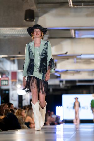 Fashion model showcases mint green outfit with black vest, fringe skirt and white boots on catwalk.
