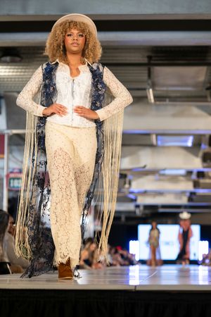 Model wears white lace ensemble with fringed navy overlay on catwalk.
