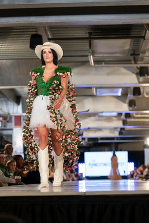 Model wearing white outfit with floral accents and wide-brimmed hat walks fashion runway.
