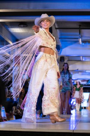 Model in flowing white lace dress with dramatic fringe details walks runway under blue lighting.