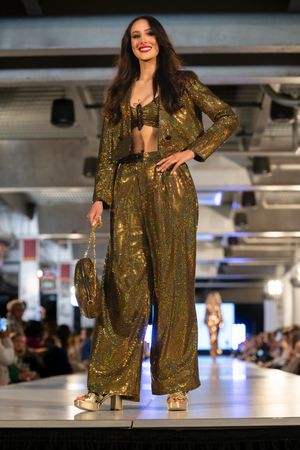 Model struts down catwalk in shimmering gold sequined pantsuit during fashion show.