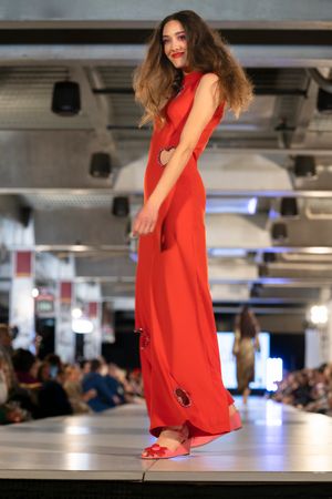 Model walks runway in flowing red sleeveless dress during fashion show with dramatic lighting.