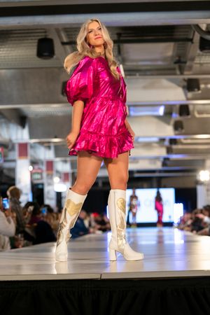 Model walks runway in hot pink ruffled dress and white cowboy boots during fashion show.