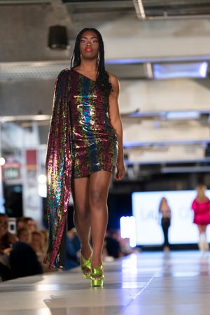 Sequined rainbow dress with asymmetrical design sparkles under runway lights during fashion show.