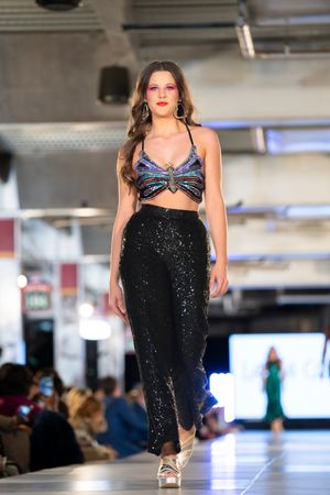 Fashion model walks runway in sparkly black evening gown and metallic heels at indoor fashion show.