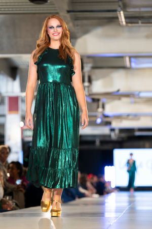 Model showcases emerald green metallic babydoll dress with gold boots on indoor catwalk.