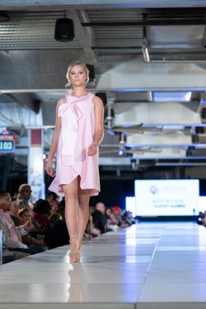 Model walks runway in pink cold-shoulder dress at fashion show with blue lighting and audience in background.