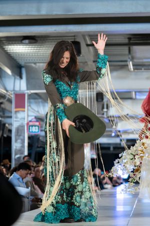 Model in emerald green ensemble with fringe details performs dramatic pose on catwalk.
