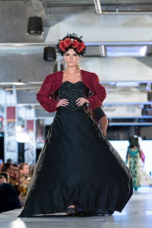 Dramatic black ball gown with burgundy accents and floral headpiece featured on indoor catwalk.