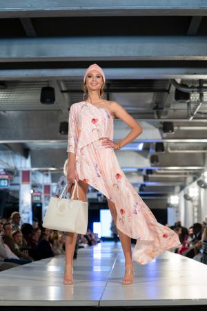 Model presents light pink floral dress with white handbag on fashion runway.
