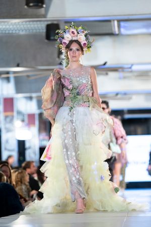 Model showcases ethereal white gown with floral details and elaborate floral headpiece on illuminated runway.