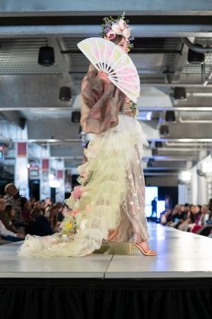 Model displays romantic pastel gown with decorative fan and floral crown during fashion show performance.
