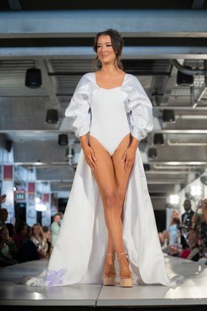 Fashion model in flowing white dress walks down illuminated runway during catwalk show.