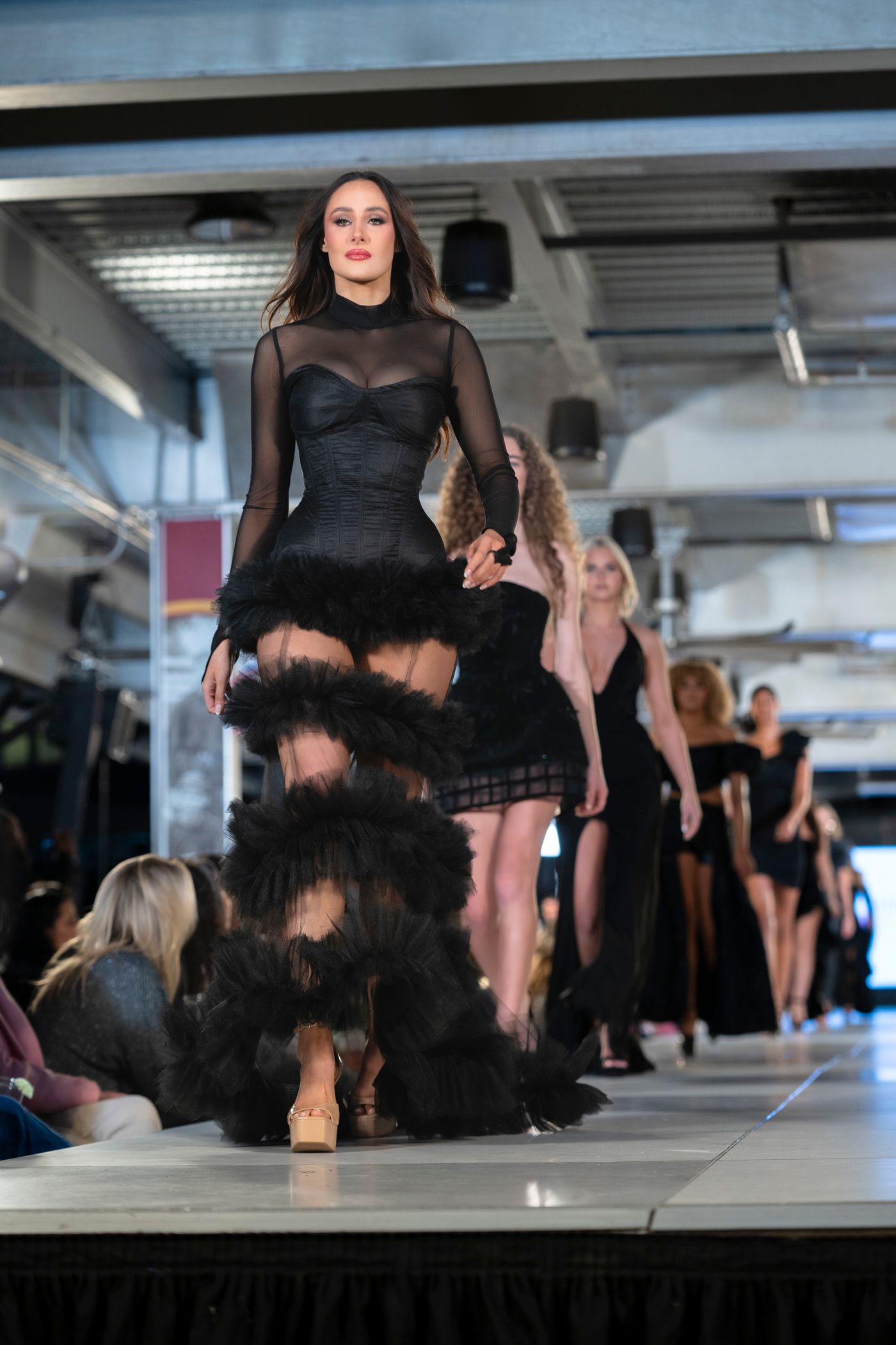 Dramatic black sheer gown with feather details displayed on illuminated fashion runway.