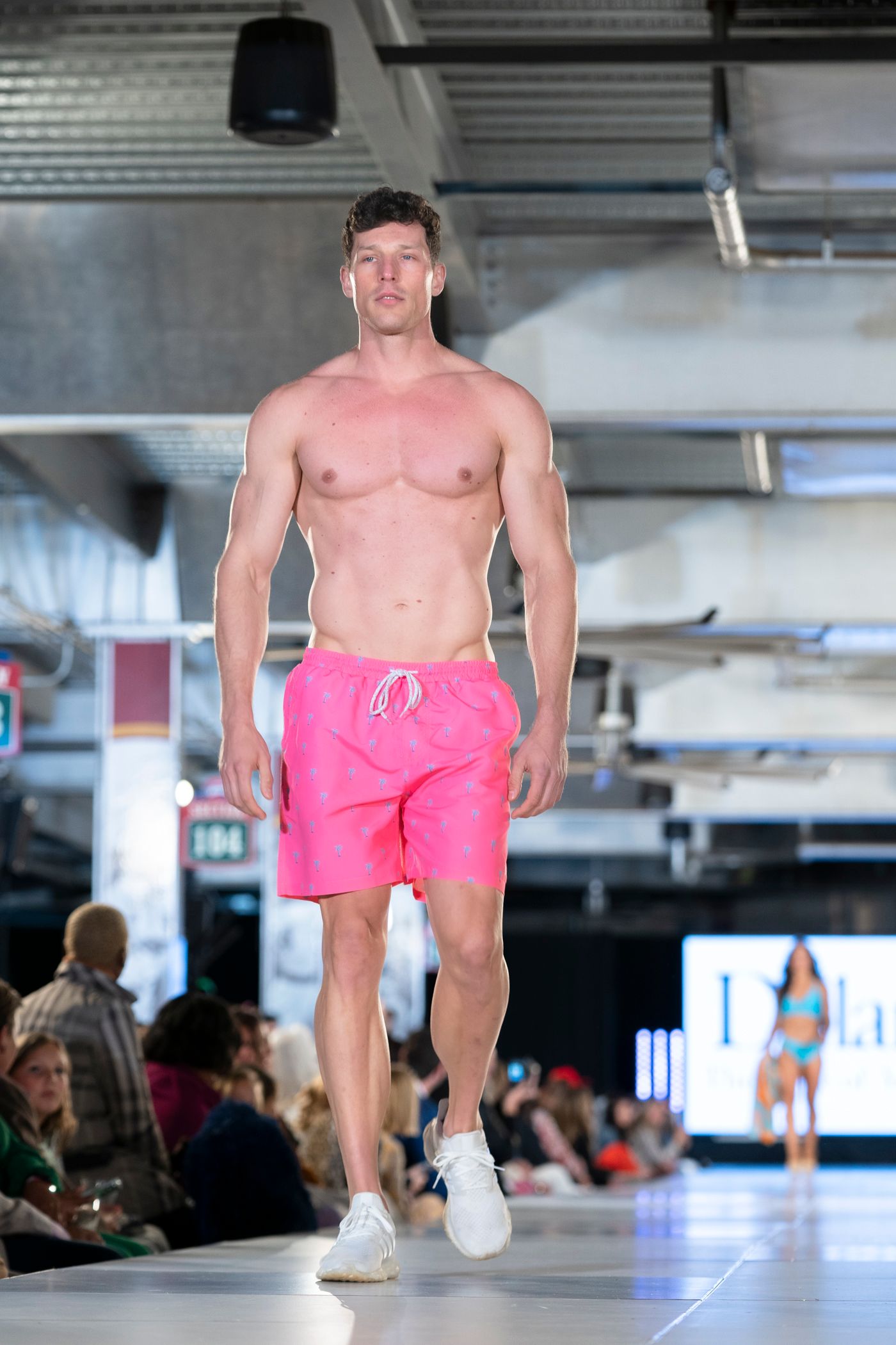 Model showcases bright pink swim shorts on a fashion runway with white sneakers.