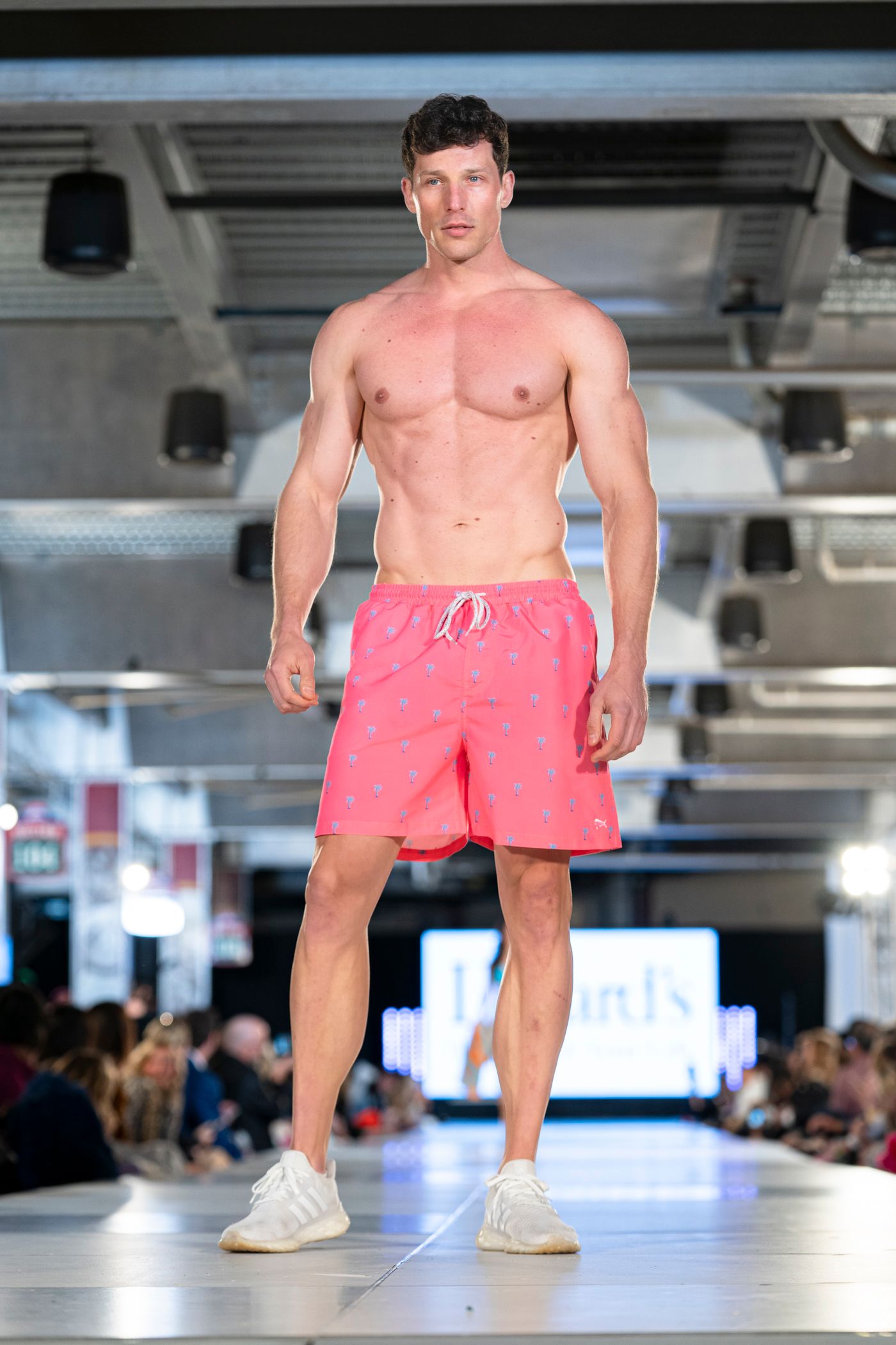 Model walks runway in pink swim shorts and white sneakers during summer fashion show.