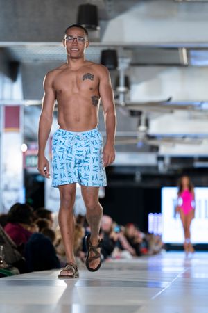 Model showcases light blue patterned swim shorts on runway during beachwear fashion show.