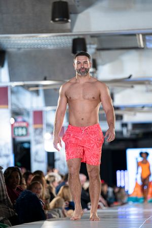 Model walks runway in coral red swim shorts at indoor fashion show.