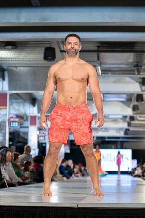 Model showcases coral patterned swim trunks on illuminated runway with audience in background.