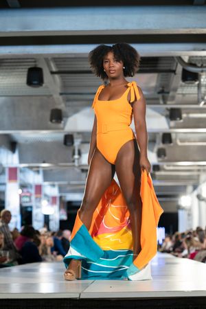Model walks runway in vibrant orange and turquoise color-blocked swimwear at fashion show.